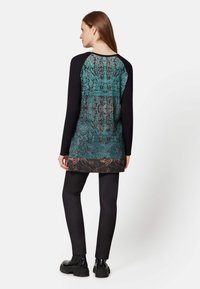 Schwarzes Langarmshirt mit einem türkis-schwarz gemusterten Rücken, ausgestattet mit einem geschwungenen Saum und leichtem Stoff, kombiniert mit schwarzen Leggings.