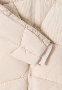 Beige polstret jakke med kviltede ærmer og en lodret lynlås, der har ribbet stof på overkroppen og manchetterne.