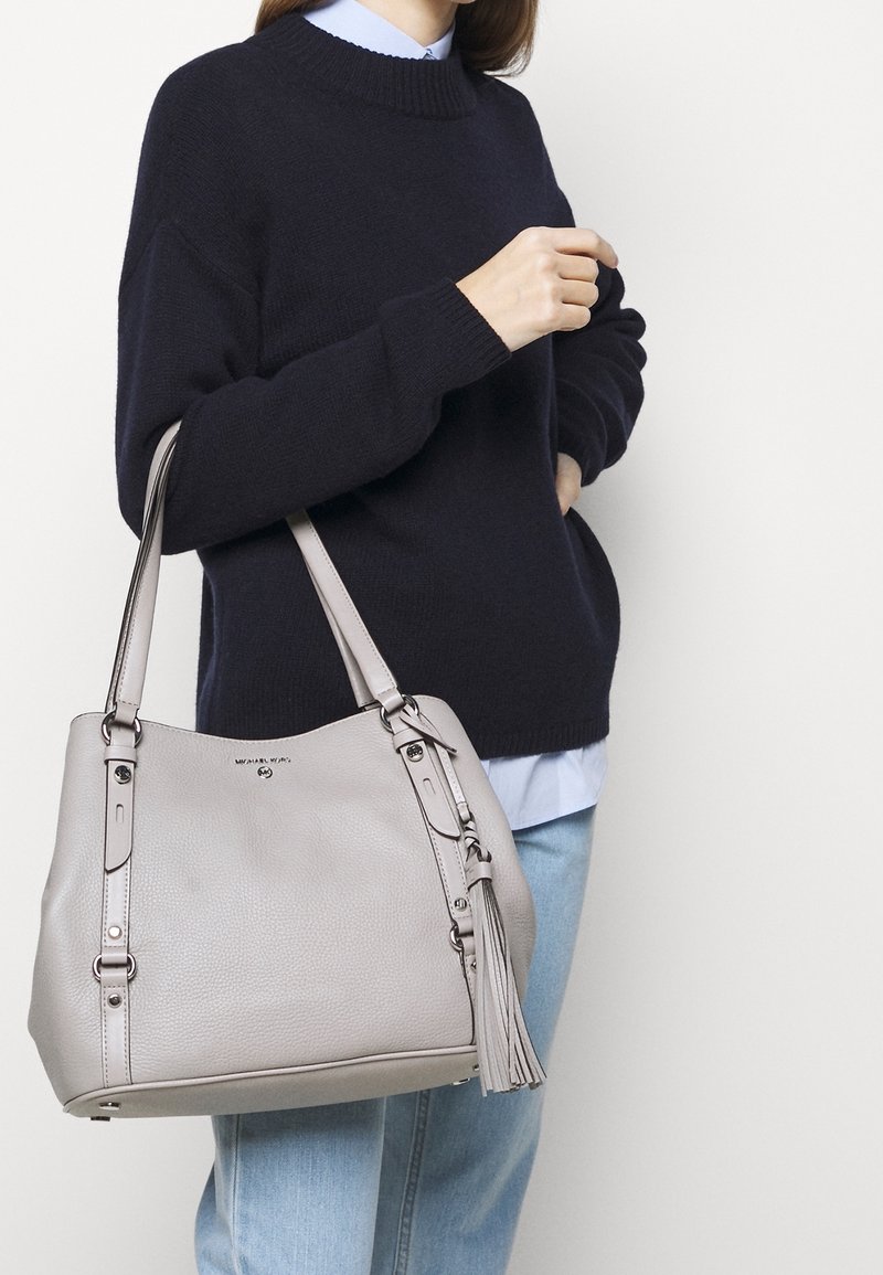 Lysegrå læder tote bag med tekstureret overflade, dobbelte hanke og dekorative frynsedetaljer. Båret med en marineblå sweater og en blå skjorte.