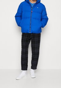 Niebieska kurtka puchowa z zamkiem z przodu i logo. Zestawiona z czarnymi spodniami w kratę i białymi trampkami. Gładka tekstura, izolowany design.