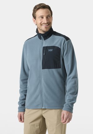 Hombre sonriendo, vestido con chaqueta de forro polar azul claro con cremallera y bolsillo en el pecho azul oscuro, y pantalones beige, de pie frente a un fondo liso.
