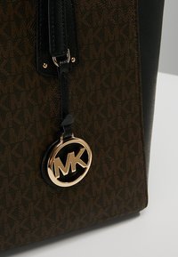 Bruine gestippelde tote bag met zwarte accenten, voorzien van een gouden MK-logo charm en textuurstof. Ritssluiting aan de bovenkant.