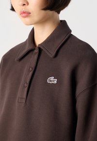 Dark brown polo shirt in soft fabric featuring a classic collar, three buttons, and a white embroidered crocodile logo on the left chest.