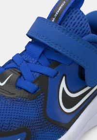 Kék sportcipő, hálós felsőrésszel, fekete részletekkel, állítható pánttal és fehér Nike pipa logóval az oldalon.