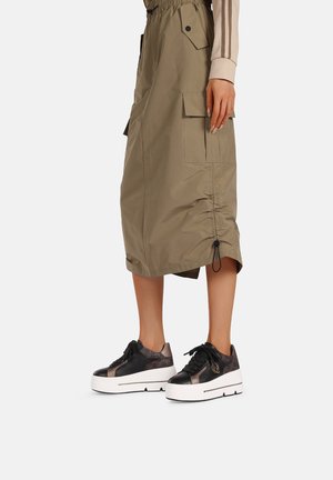 Gonna midi khaki con fascia elastica in vita e tasche laterali, abbinata a sneakers nere e metalliche con suola bianca spessa e design con lacci.