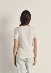 Mujer con cabello largo y castaño de pie mirando hacia la pared, lleva una camiseta blanca de manga corta y jeans azul claro.