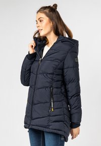 Jonge vrouw in een marineblauwe gewatteerde capuchonjack, die de kraagrits vasthoudt, staande tegen een effen lichte achtergrond.