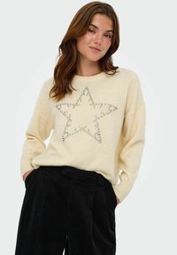 Crème trui met een grote zilveren sterdesign van pailletten op de voorkant. Heeft geribbelde manchetten en een relaxte pasvorm. Zwarte corduroy broek.