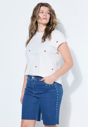 Kvinde iført hvid T-shirt med små røde hjerter og blå denimshorts med sølvnitter, står med den ene hånd i lommen.