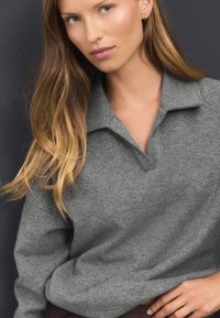 Polo gris en tricot avec un col et une coupe légèrement décontractée. La texture est lisse avec une finition mate. Le vêtement est porté avec des cheveux longs.