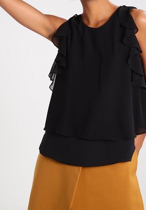 Femme portant un haut noir sans manches à volants et un pantalon jaune moutarde, un bras levé sur un fond blanc uni.