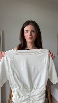 Bela kratkotrajna majica s kratkimi rokavi in nabranim pasom, izdelana iz gladkega materiala. Ima okroglo izreza in rahlo sijoč videz, prikazana na modelu.