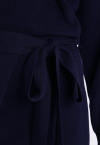 Tessuto a maglia blu navy con finitura testurizzata, dotato di una vita legata. Il design include un dettaglio a fiocco e maniche lunghe.