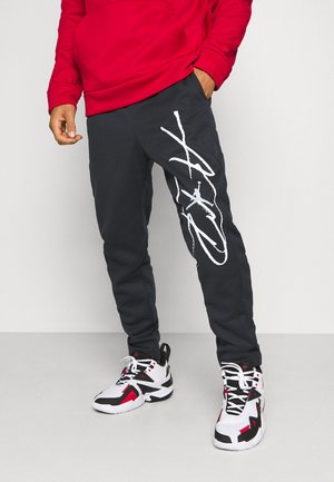 Mann in schwarzen Jogginghosen mit weißem grafischen Text auf dem linken Bein, rotem Hoodie und weißen, schwarzen und roten Sportschuhen, der an einer schlichten Wand steht.