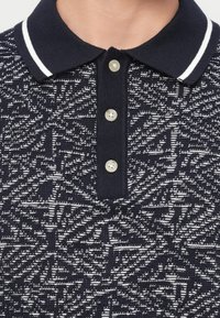 Πόλο μπλουζάκι σε ναυτικό μπλε χρώμα με λευκό γεωμετρικό πλεκτό σχέδιο, τρία λευκά κουμπιά και γιακά με λευκές ρίγες.