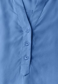 Camicia azzurra chiaro con scollo a V, tre bottoni coordinati sul davanti e una texture di tessuto morbida e liscia.