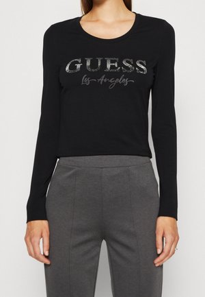 Femme portant un haut noir à manches longues avec le texte pailleté « GUESS Los Angeles » et un pantalon gris taille haute.