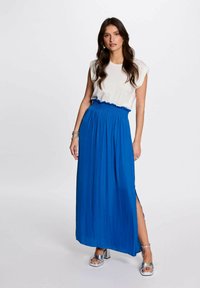 Gonna maxi in tessuto blu brillante con vita arricciata, dotata di uno spacco laterale. Indossata con una maglietta bianca e sandali con tacco argento.