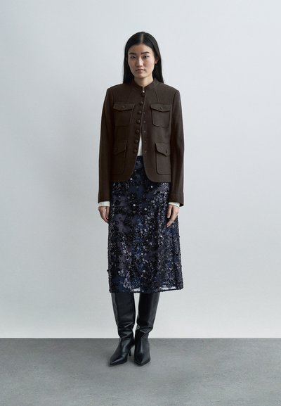 Γυναίκα που φοράει καφέ σακάκι με κουμπιά και μπροστινές τσέπες, μπλε σκούρα φούστα με παγέτες, άσπρο τοπ και μαύρες μπότες μέχρι το γόνατο, στέκεται σε γκρι πάτωμα.