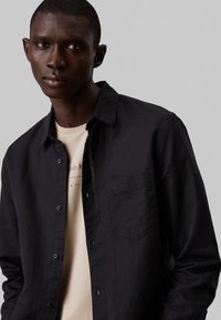 Camisa negra de botones con un bolsillo en el pecho, fabricada con un tejido texturizado, superpuesta a una camiseta beige de Calvin Klein con una marca sutil.