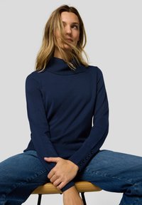 Marineblauer Rollkragenpullover aus weichem Material mit gerippten Ärmelbündchen und Knopfdetails. Kombiniert mit Jeans für einen lässigen Look.