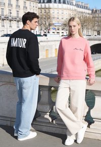 Homme en sweat-shirt noir avec le texte « AMOUR » et jean clair se tient à côté d'une femme en sweat-shirt rose avec le texte « Mira Paris » et pantalon clair au bord de la rivière.