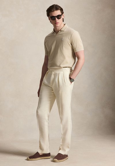 Άνδρας που φοράει μπεζ πόλο μπλούζα, κρεμ πλισέ παντελόνια, καφέ espadrilles, γυαλιά ηλίου και ρολόι χειρός, στέκεται μπροστά σε ουδέτερο φόντο.