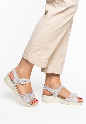 Feet wearing beige floral wedge sandals with ankle straps and silver buckles, paired with rolled-up light beige corduroy pants.