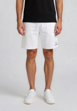 Man draagt witte trekkoordshorts met logo op linkerbeen, zwart shirt en witte sneakers, staande tegen een effen achtergrond.