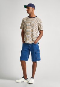 Camiseta a rayas beige y marino con cuello redondo, combinada con pantalones cortos de carga azules con bolsillos laterales. Zapatillas blancas y gorra Marines.