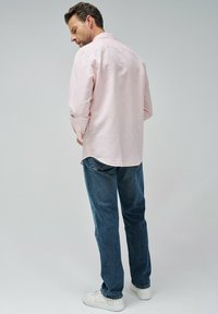 Camisa de manga larga rosa claro con cuello abotonado, combinada con jeans azules y zapatillas blancas. Presenta un corte holgado y un pliegue en la parte posterior.