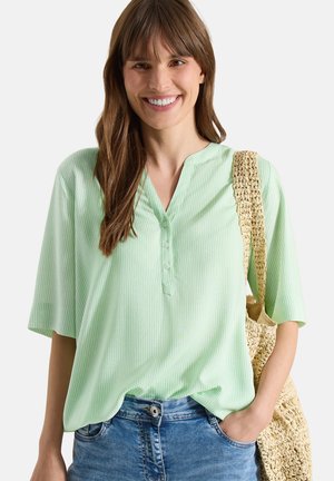 Femme souriante portant un chemisier rayé vert clair et un jean bleu, portant un sac en paille tressée sur l’épaule.