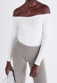 Weißes, schulterfreies Langarmoberteil, kombiniert mit gerippten, beigen High-Waist-Hosen mit vertikalen Streifen und strukturierter Oberfläche.