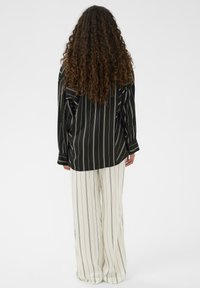 Ženska z dolgimi kodri, oblečena v črno sivo prugasto srajco in bele široke hlače s prugami, stoji obrnjena stran od fotoaparata na belem ozadju.