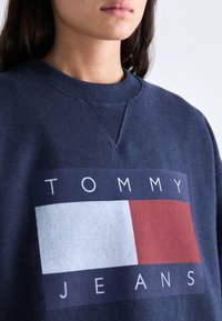 Námornícka mikina s okrúhlym výstrihom a rebrovanými manžetami, má obdĺžnikový grafický motív v modrej, bielej a červenej farbe s textom "TOMMY JEANS".