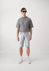Grå kortermet hettegenser og lyseblå slitte denimshorts. Modellen har på seg svarte solbriller og hvite joggesko med grå sokker.