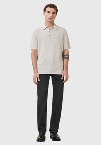 Beige polo-shirt met korte mouwen van een gestructureerde stof, drie-knopen sluiting en een contrasterende kraag, gedragen met zwarte slim-fit jeans en zwarte schoenen.