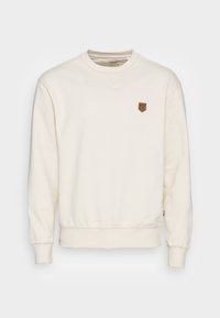 Krémová mikina s ribbovaným kulatým výstřihem, dlouhými rukávy a předním hrudním nášivkem ze strukturovaného hnědého materiálu.