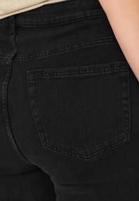Jeans de mezclilla negra con una textura suave; presentan un bolsillo trasero y costuras en tono. El tejido parece duradero y ligeramente elástico.