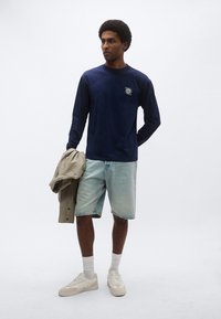 Μπλε σκούρο πουκάμισο με μακριά μανίκια και γραφικό του ήλιου; ανοιχτό μπλε τζιν σορτς; μπεζ μπουφάν; κρεμ αθλητικά παπούτσια; υφές υφασμάτων; καθημερινή ενδυμασία.