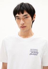 Witte katoenen t-shirt met een tekstafbeelding in het blauw die "NIETS VERANDERT" op het linkerborstgebied zegt. Korte mouwen en een ronde halsontwerp.