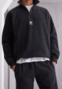 Svart fleece-pullover med halv dragkedja i kragen, med en vit Adidas-logga på bröstet och ribbade muddar. Matchas med mörka sweatpants.