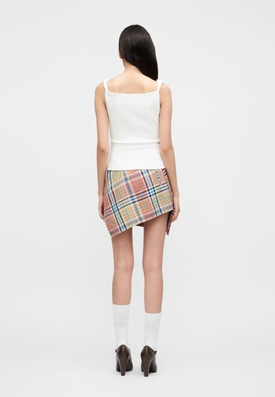 Γυναίκα που στέκεται με την πλάτη προς το θεατή, φοράει λευκή αμάνικη μπλούζα, ασύμμετρη πολύχρωμη μίνι φούστα καρό, λευκές κάλτσες και καφέ παπούτσια με ψηλό τακούνι.