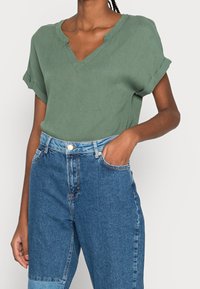 Blusa verde a maniche corte con scollo a V e maniche arrotolate, abbinata a jeans in denim blu a vita alta con una leggera macchia su una gamba.