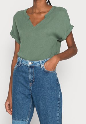 Blusa verde a maniche corte con scollo a V e maniche arrotolate, abbinata a jeans in denim blu a vita alta con una leggera macchia su una gamba.