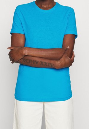 Henkilö, joka pukeutunut kirkkaan siniseen lyhythihaiseen paitaan ja valkoisiin housuihin, käsivarret ristissä, vasemmassa kyynärvarressa näkyy roomalainen numerotatuointi.