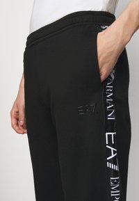 Czarne dresy z elastycznym paskiem w talii, boczną kieszenią, wytłoczonym logo EA7 oraz białym nadrukiem marki wzdłuż boku dla kontrastu.