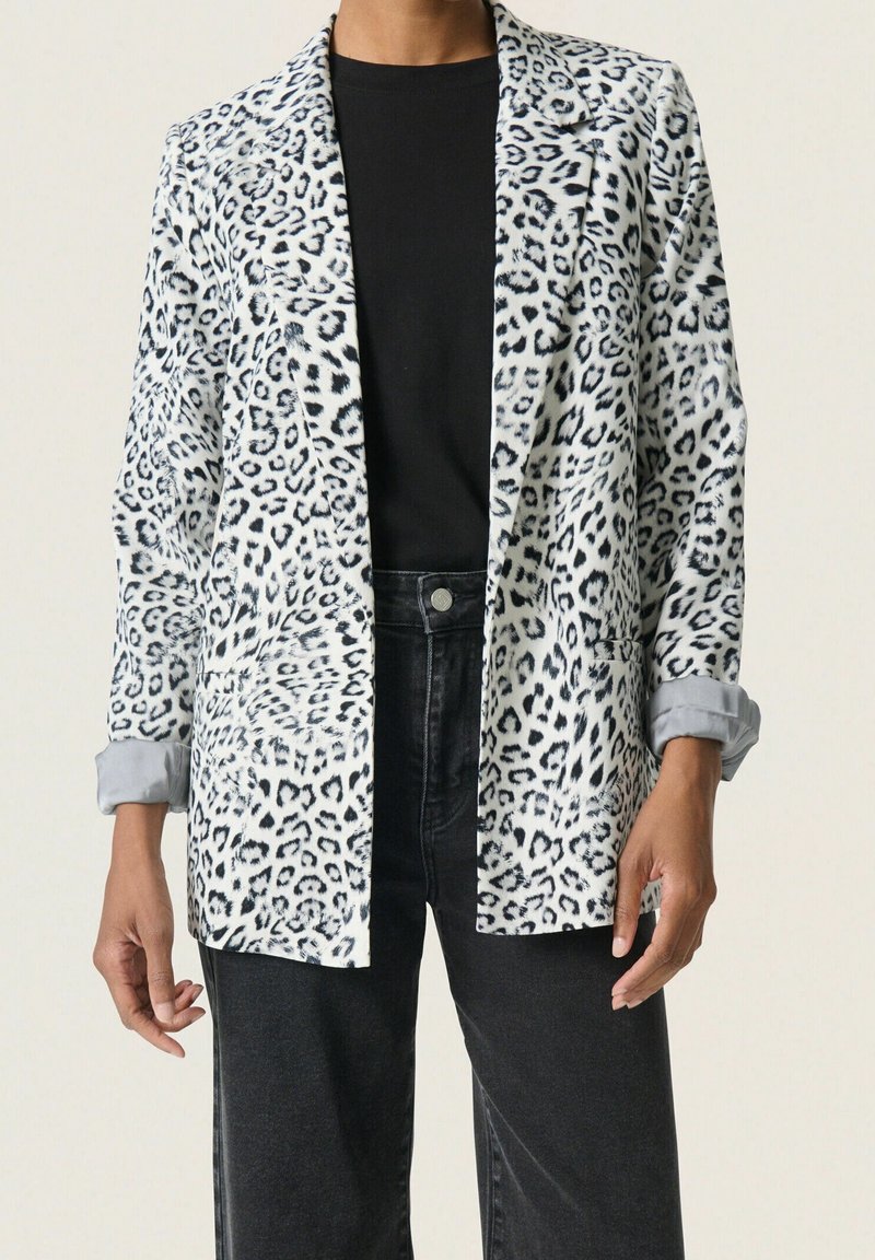 Femme portant un blazer blanc et noir à imprimé léopard sur un top noir et un jean noir taille haute, debout devant un fond clair.