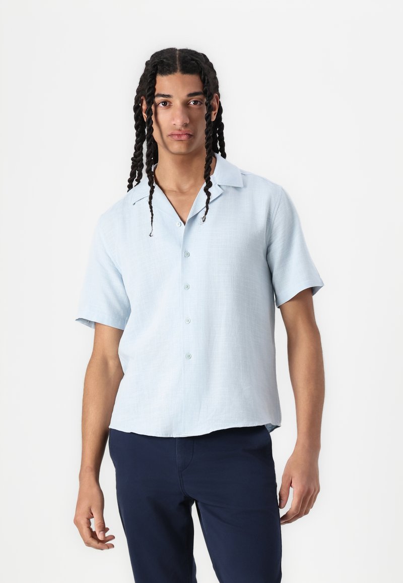 Chemise bleu clair à manches courtes avec une finition texturée, fermée par des boutons sur le devant, et une coupe décontractée, associée à un pantalon marine.