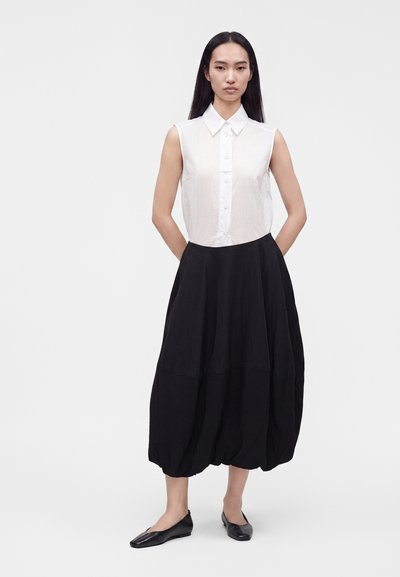 Femme debout portant un chemisier blanc sans manches à boutons, une jupe midi noire volumineuse et des ballerines noires sur fond blanc uni.